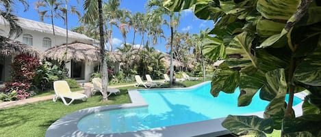 A swimming pool surrounded by palm trees and lounge chairs, with a thatched-roof building in the background.