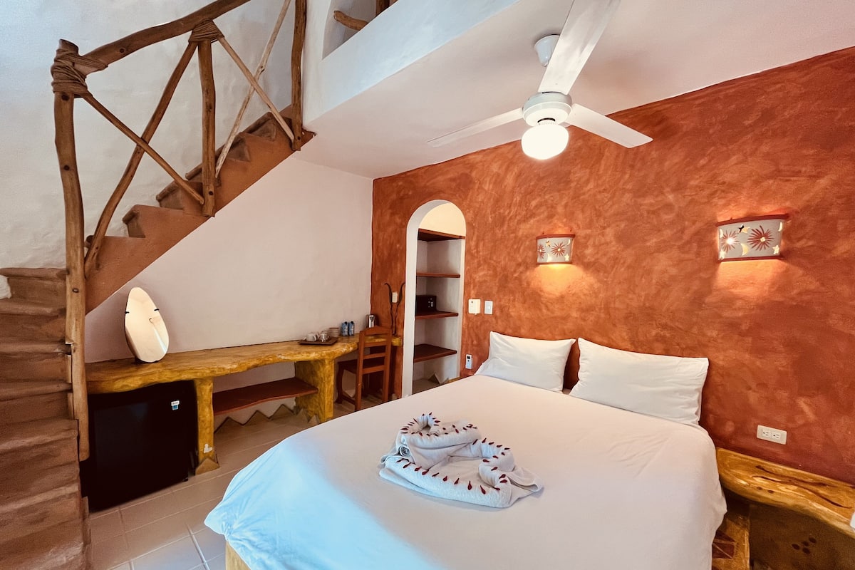 A bedroom with a bed, a desk, a chair, a ceiling fan, and a staircase.