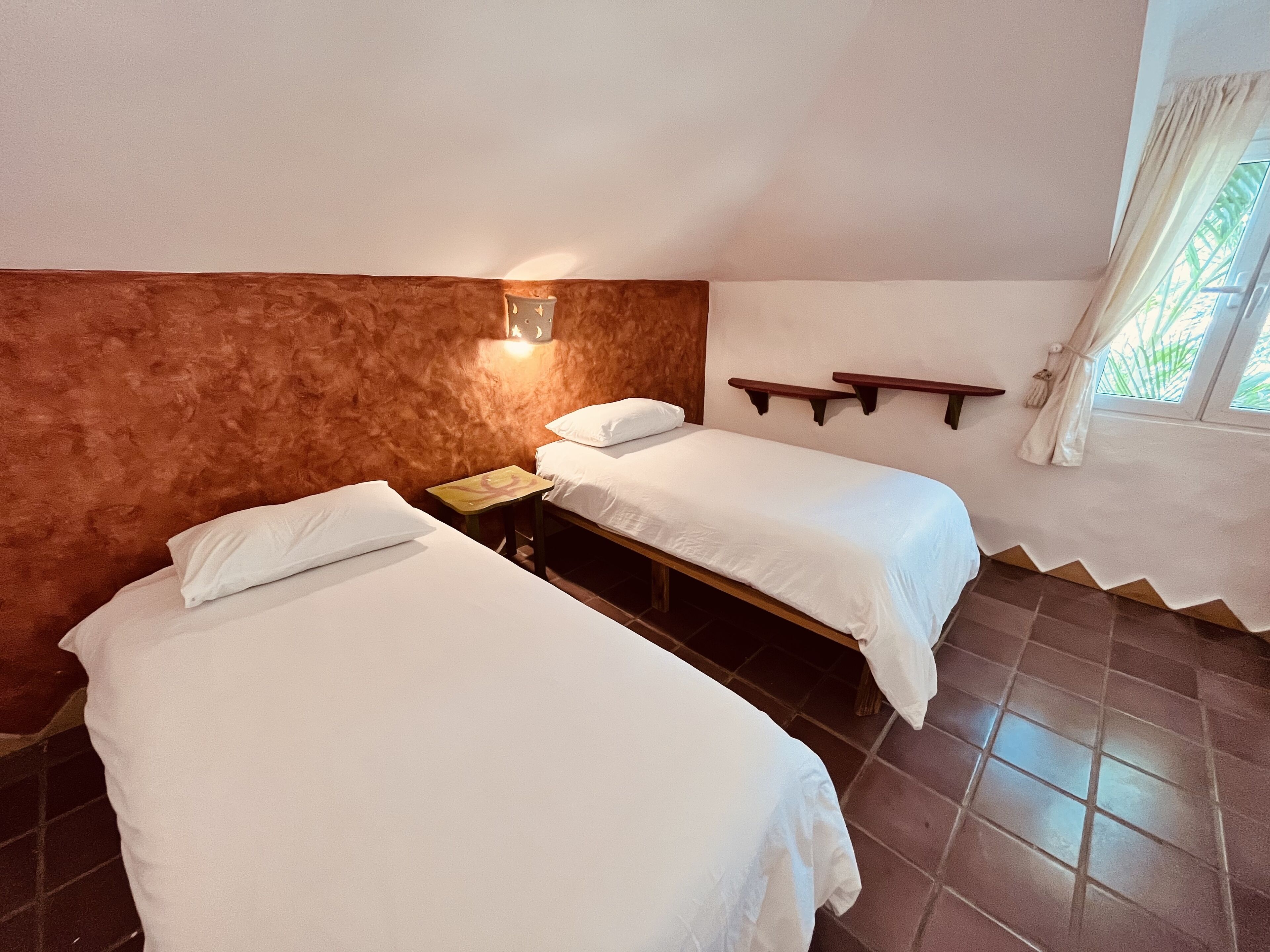 Ein Zimmer mit zwei Betten, einem Nachttisch, einer Wandablage und einem Fenster mit Vorhängen.