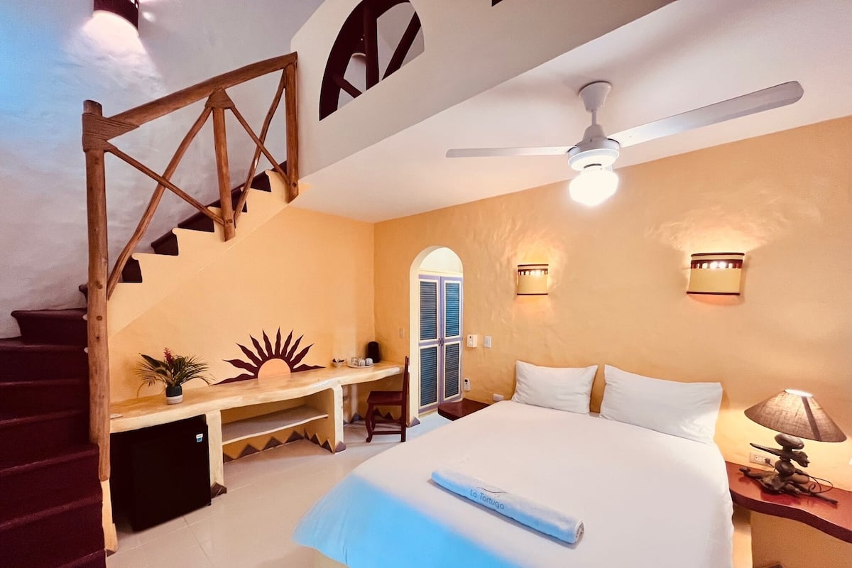 A bedroom with a bed, a desk, a chair, a staircase, and a ceiling fan.