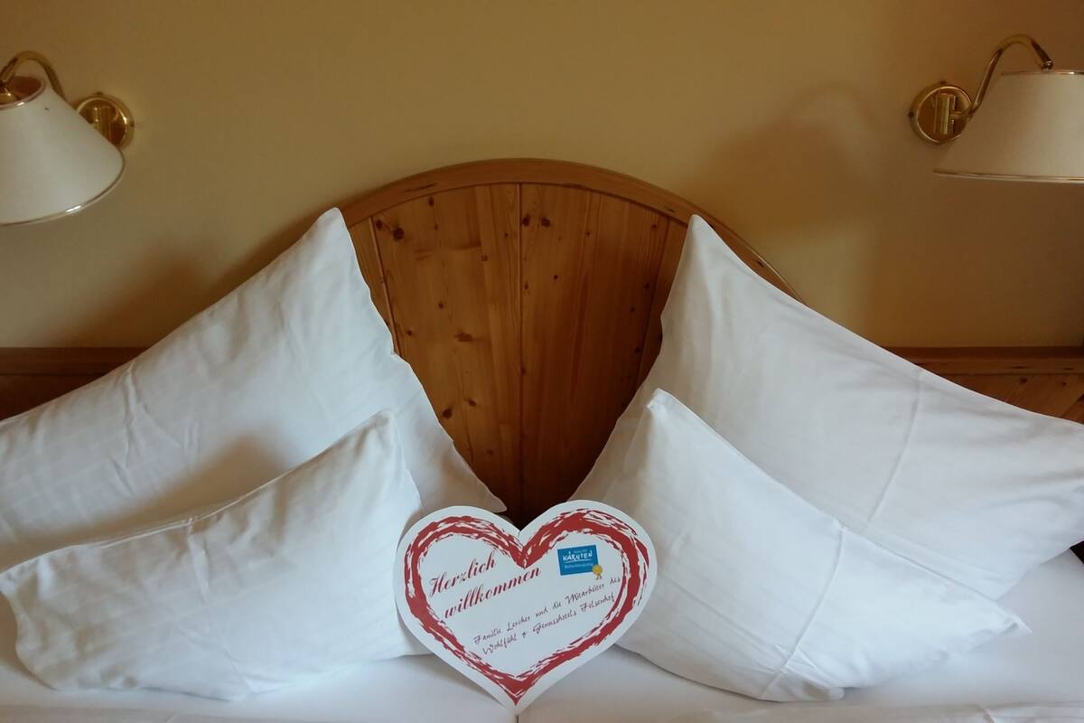 A bed with white pillows and a heart-shaped sign with text in German.