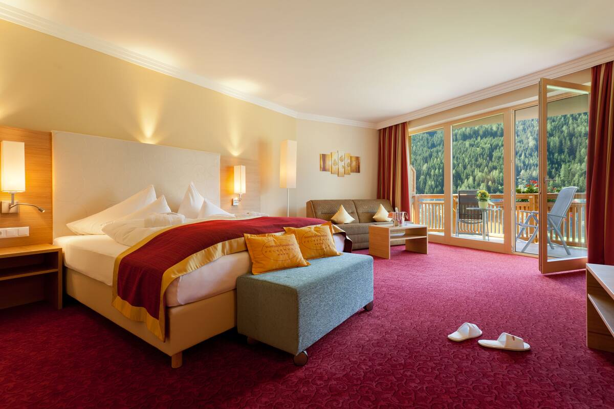 A hotel room with a large bed, a sofa, a small table, and a balcony with a view of the forest.