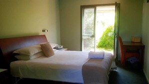 A neatly made bed with pillows and a blanket, a bedside table with a lamp, and a chair in a room with a window and a door. - Red Brier Cottage Accommodation (Richmond)