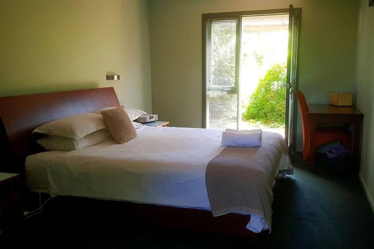 A neatly made bed with pillows and a blanket, a bedside table with a lamp, and a chair in a room with a window and a door.