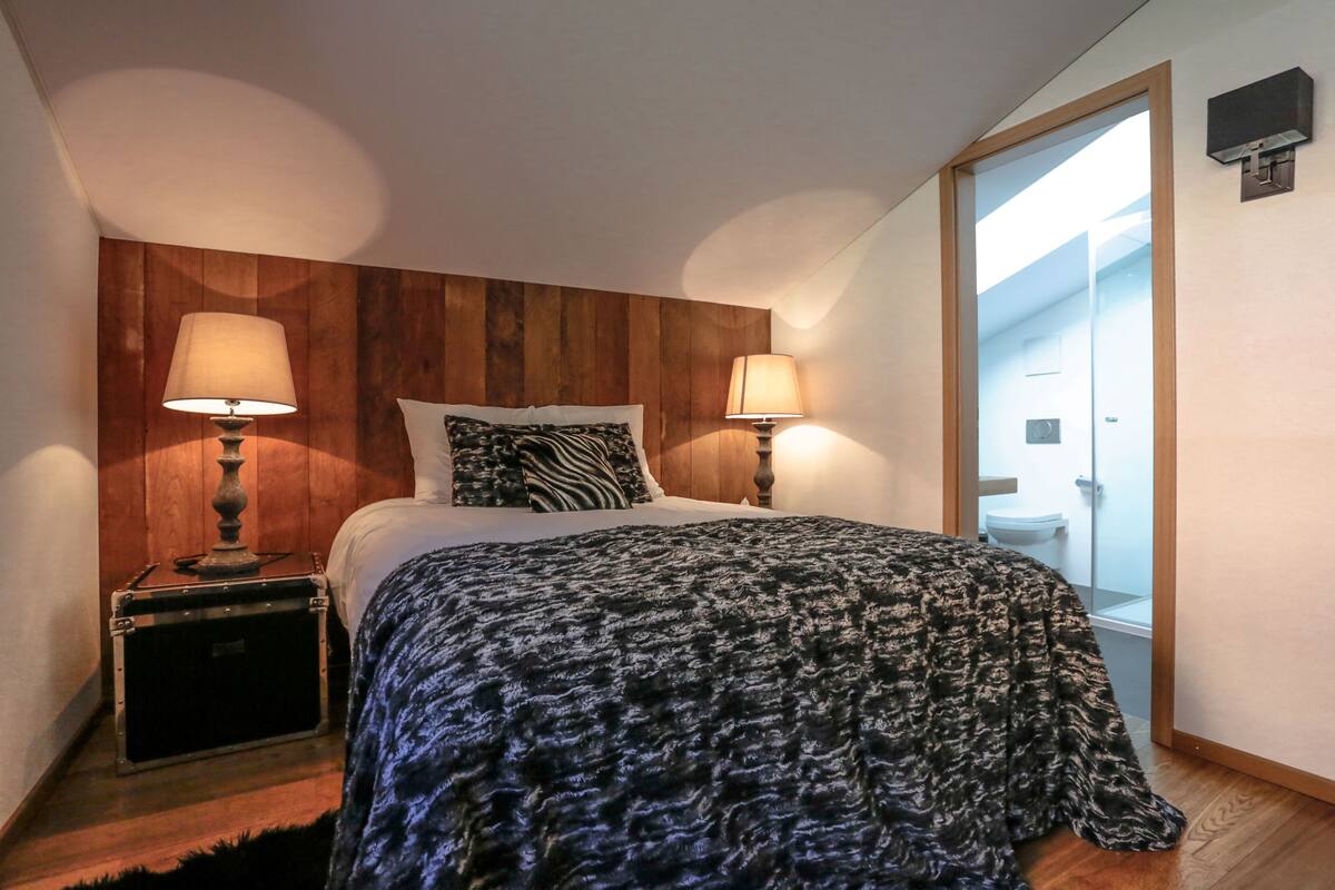 Une chambre à coucher avec une tête de lit en bois, deux lampes de chevet, un plaid à motifs zébrés et une valise posée sur la table de chevet.