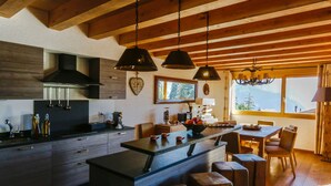 Une cuisine moderne avec des poutres en bois, des luminaires suspendus, un grand îlot central et une vue sur les montagnes par une fenêtre.