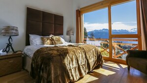 Une chambre avec un grand lit, une table de chevet, une lampe et une vue sur les montagnes par une grande fenêtre.