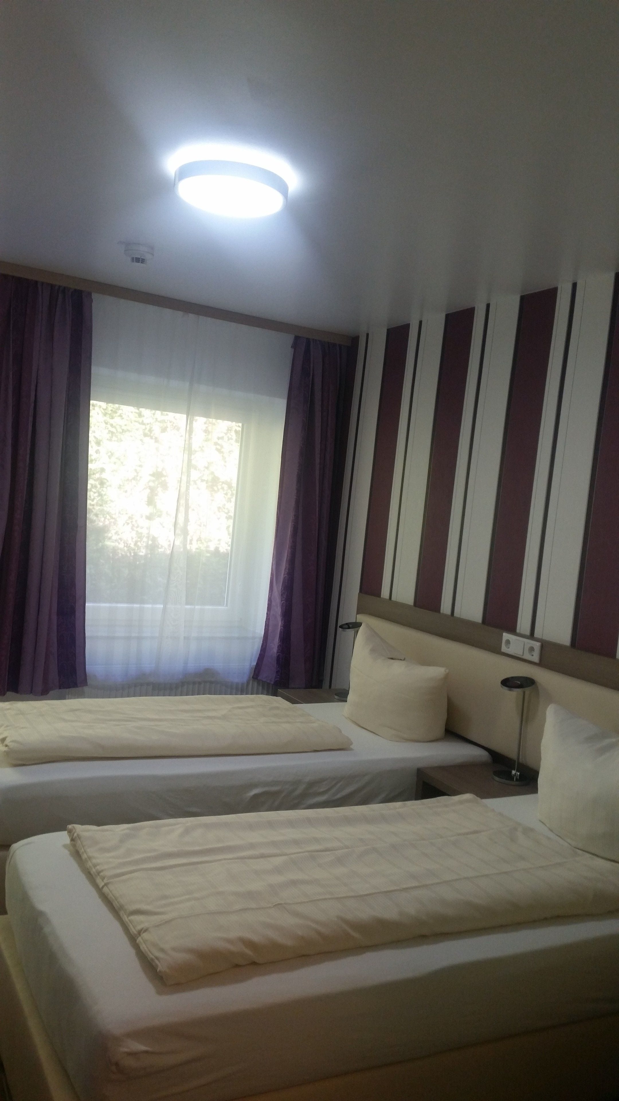 Ein Hotelzimmer mit zwei Betten, einem Fenster mit Vorhängen, einer gestreiften Wand und einer Deckenleuchte.