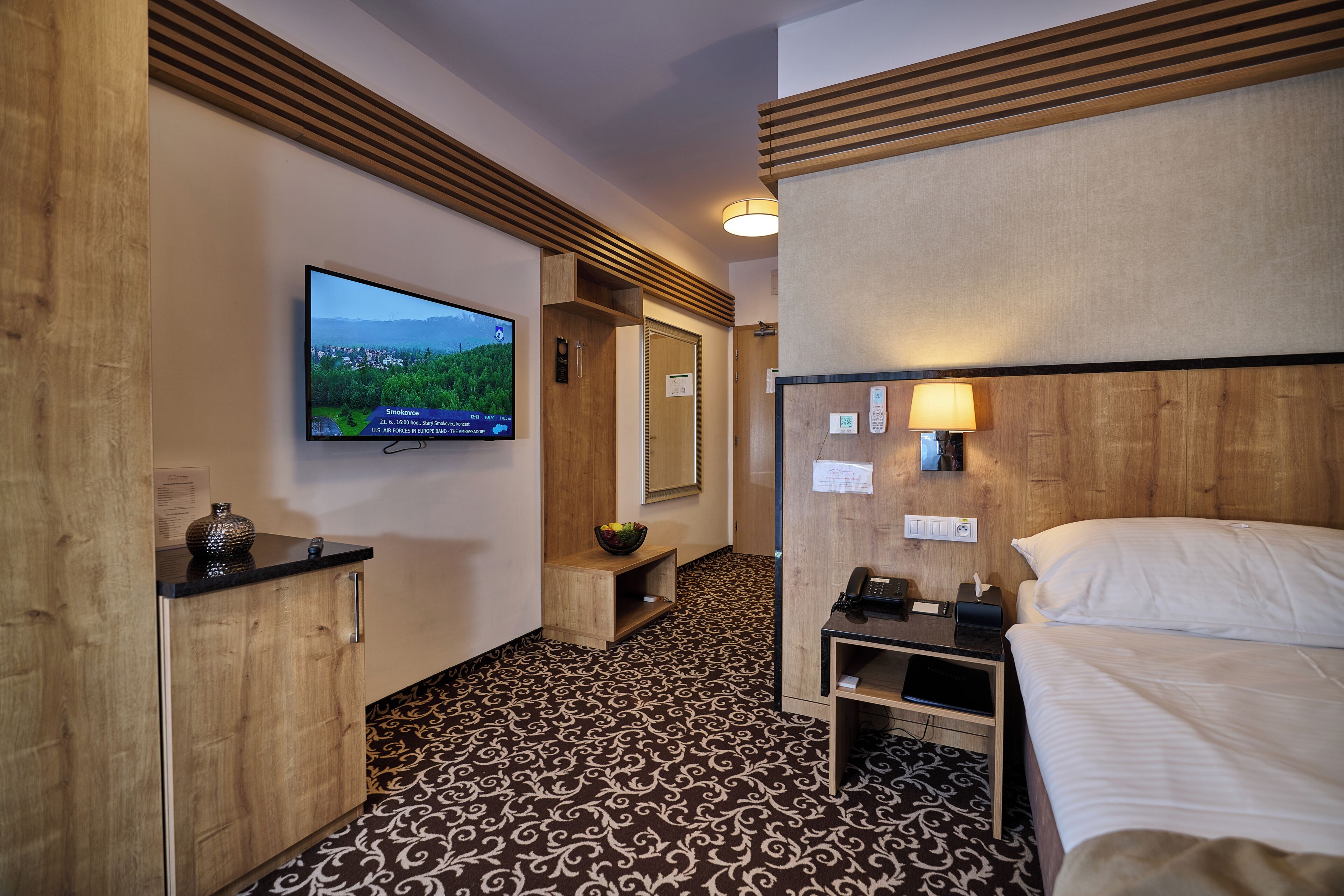 A hotel room with a bed, a television mounted on the wall, a wooden cabinet, and a patterned carpet.