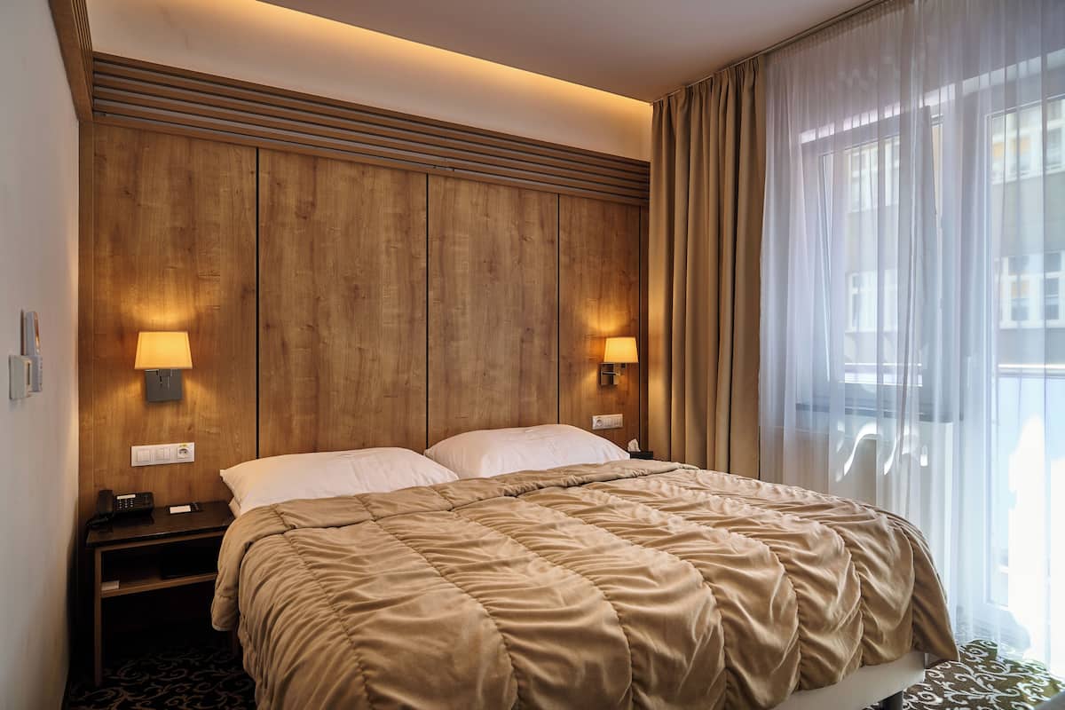 A hotel room with a wooden headboard, two bedside lamps, a bed with a beige comforter, and a window with curtains.