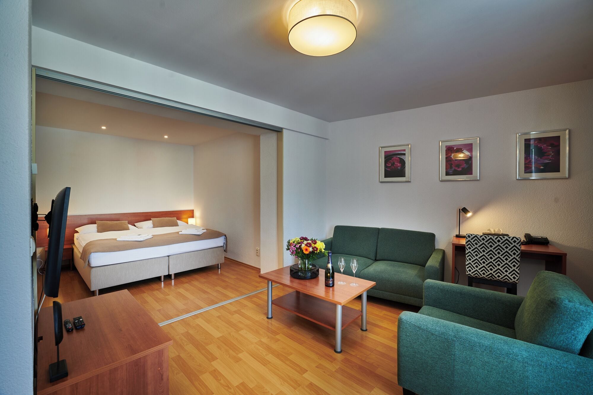 A modern hotel room with a bed, a green sofa, a wooden coffee table, and a small desk with a lamp.