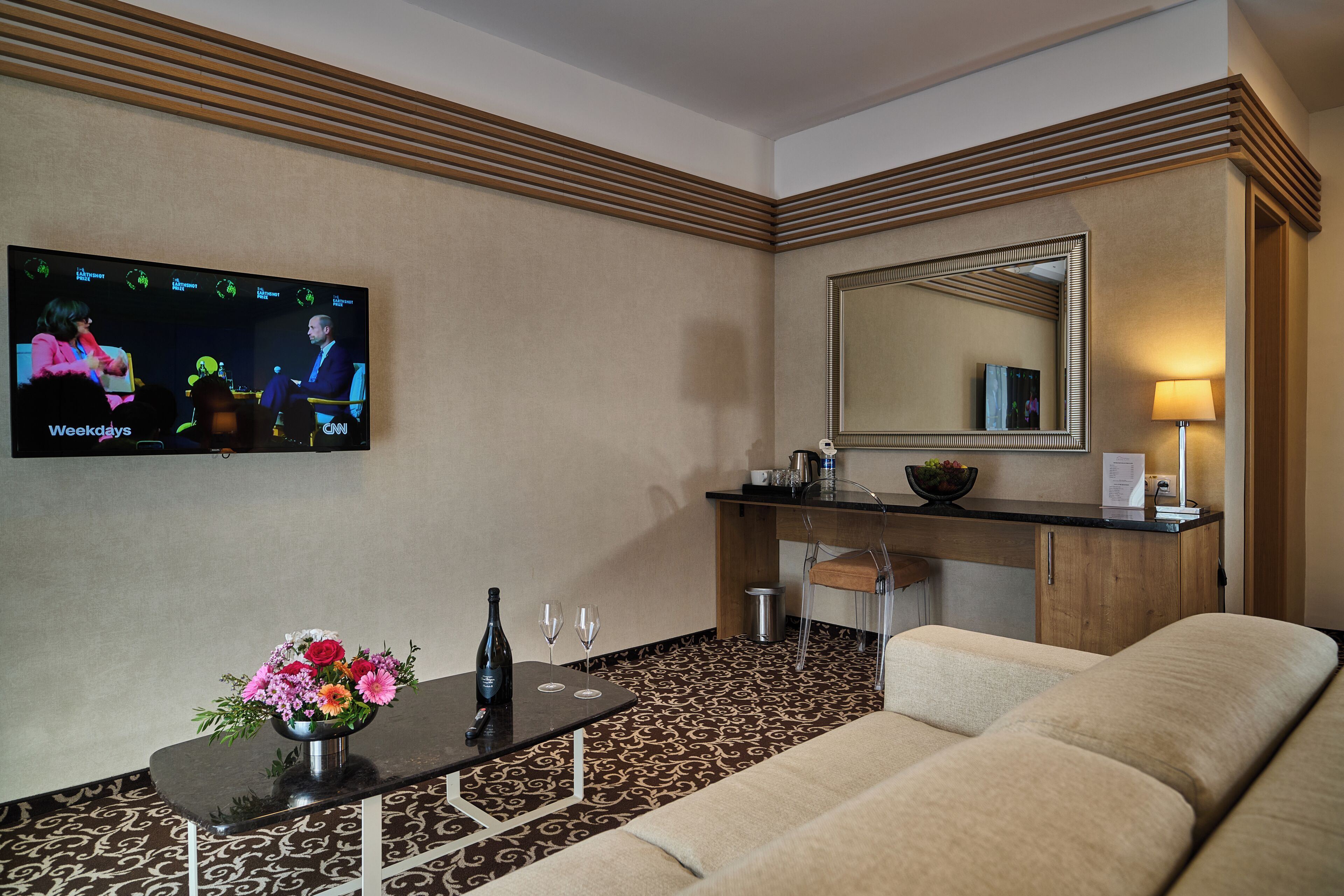 Habitación de hotel con una televisión mostrando un noticiero, un sofá, una pequeña mesa con un jarrón de flores, un espejo y un escritorio con una lámpara.