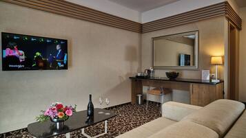 Habitación de hotel con una televisión mostrando un noticiero, un sofá, una pequeña mesa con un jarrón de flores, un espejo y un escritorio con una lámpara.