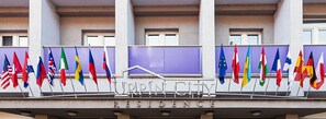 Un edificio con varias banderas, incluyendo la bandera de la Unión Europea, y las palabras "UPIN CITY RESIDENCE" visibles en la fachada.
