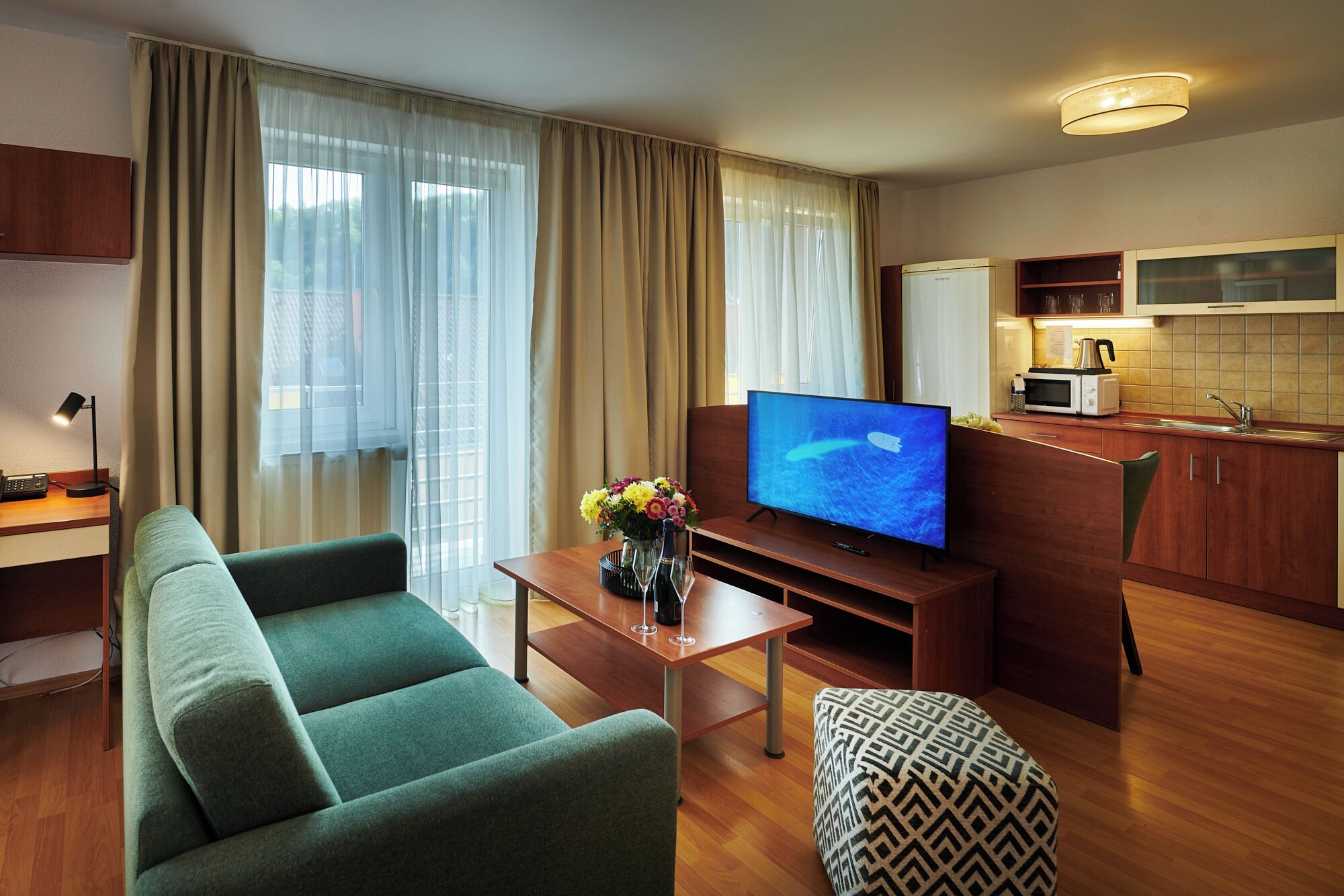 Ein modernes Hotelzimmer mit einem grünen Sofa, einem Flachbildfernseher, einem kleinen Esstisch mit Blumenvase, einer Küchenzeile und großen Fenstern mit durchsichtigen Vorhängen.
