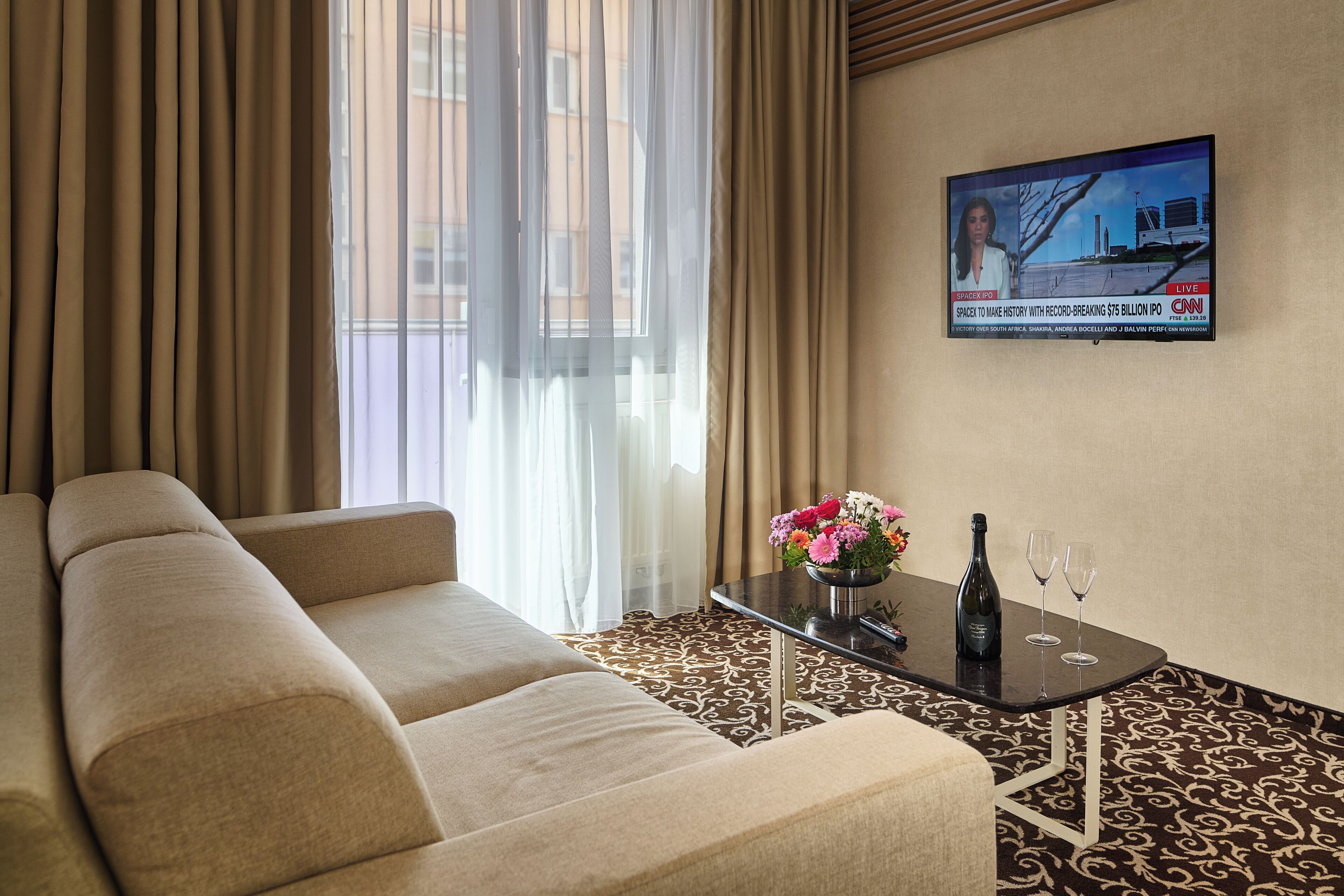 A hotel room with a sofa, a table with a bottle and glasses, curtains, and a CNN poster.
