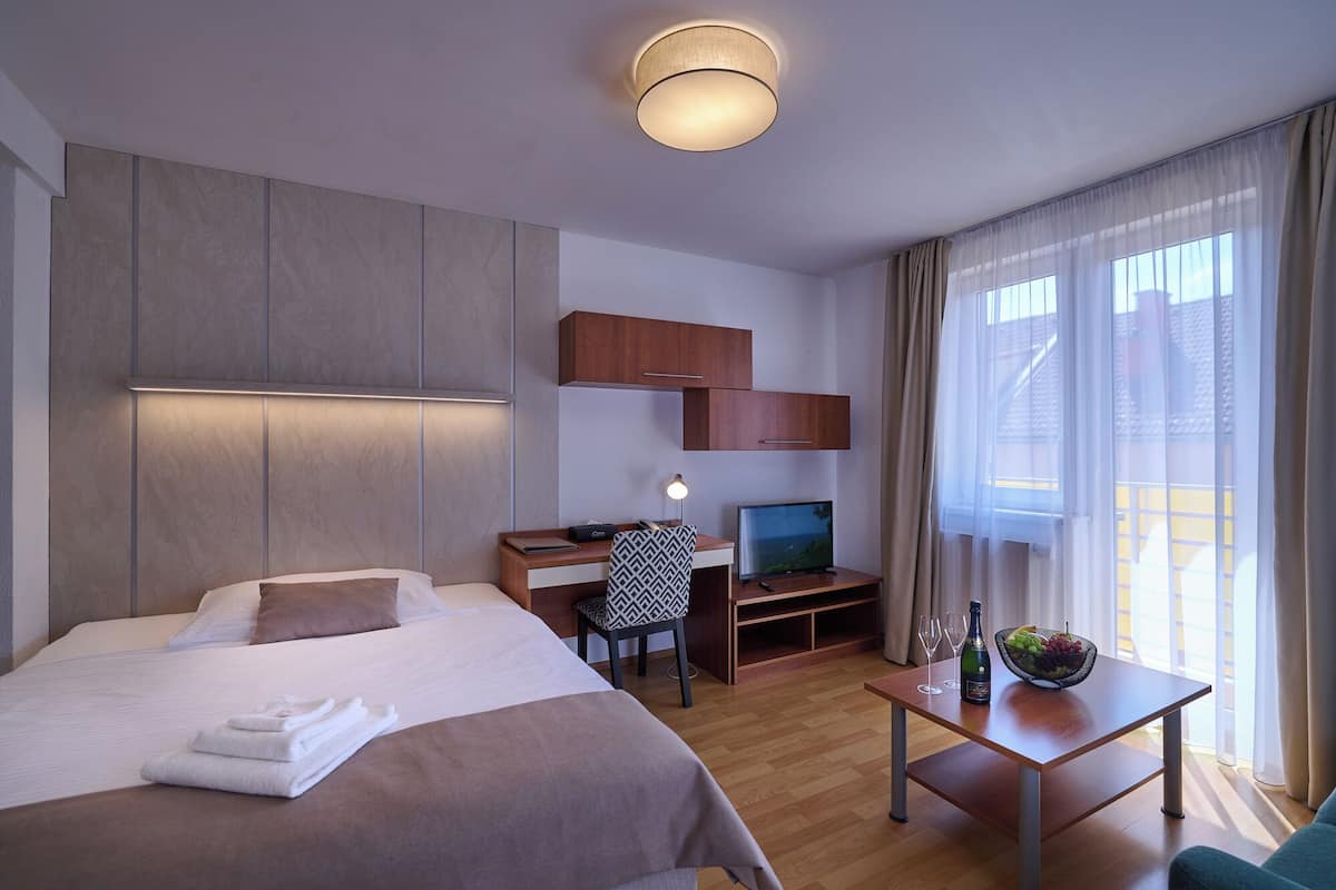 A hotel room with a bed, a desk with a computer, a TV, a small table with a bottle and glasses, and a window with curtains.
