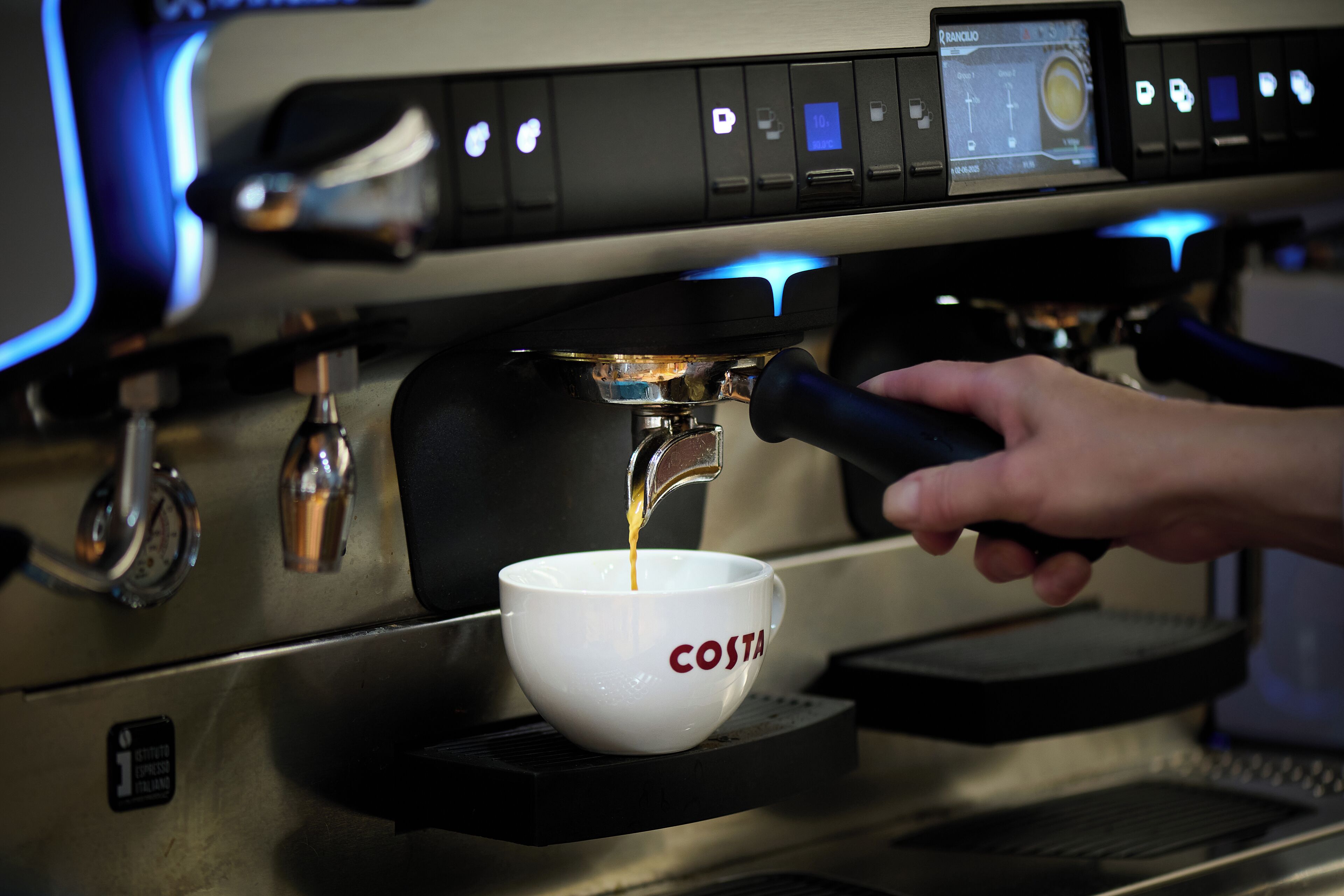 A hand operating a coffee machine, with a cup labeled "COSTA" underneath.