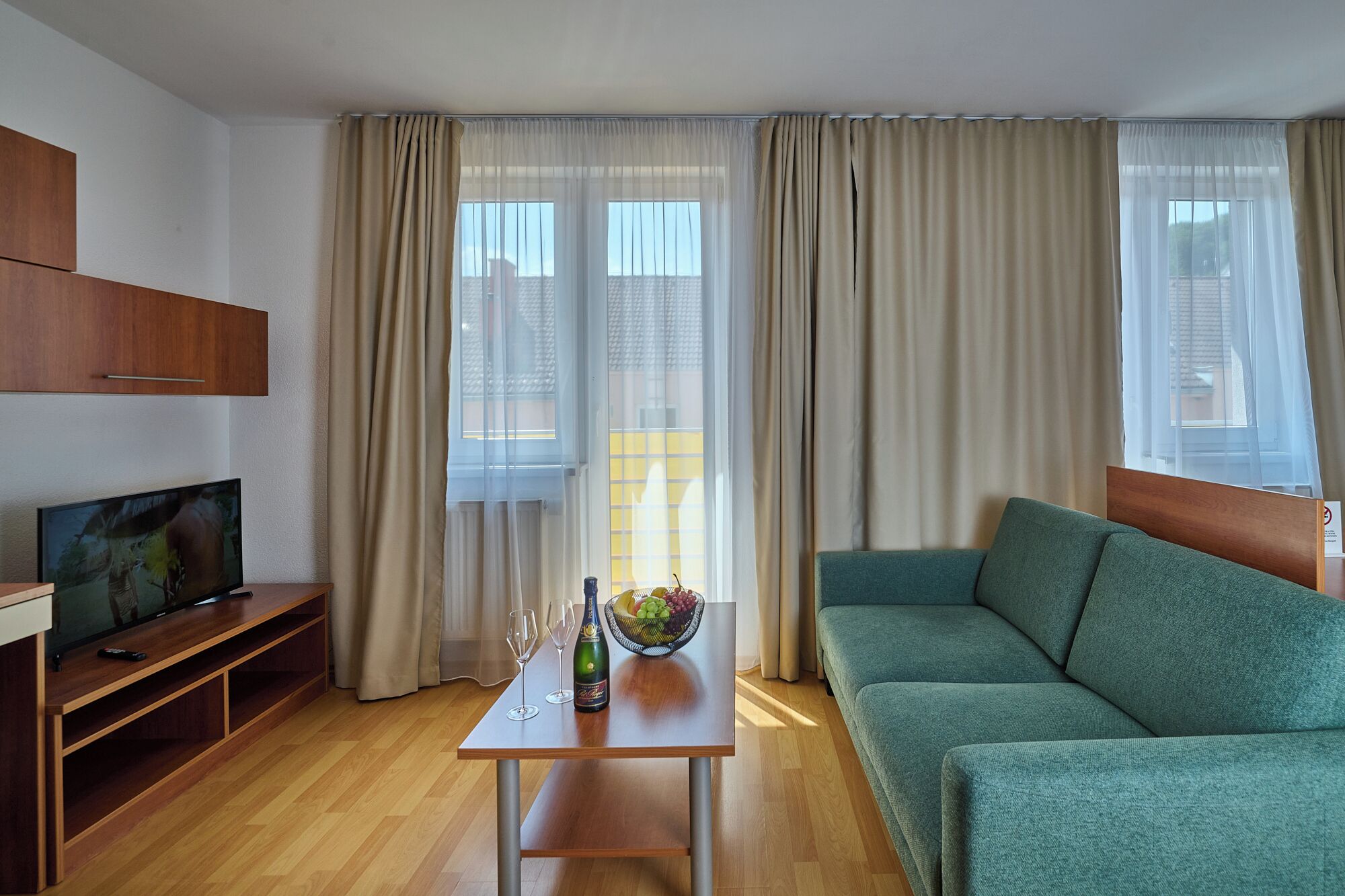 A modern living room with a green sofa, a wooden TV stand, a small table with a bottle and glasses, and large windows with sheer curtains.