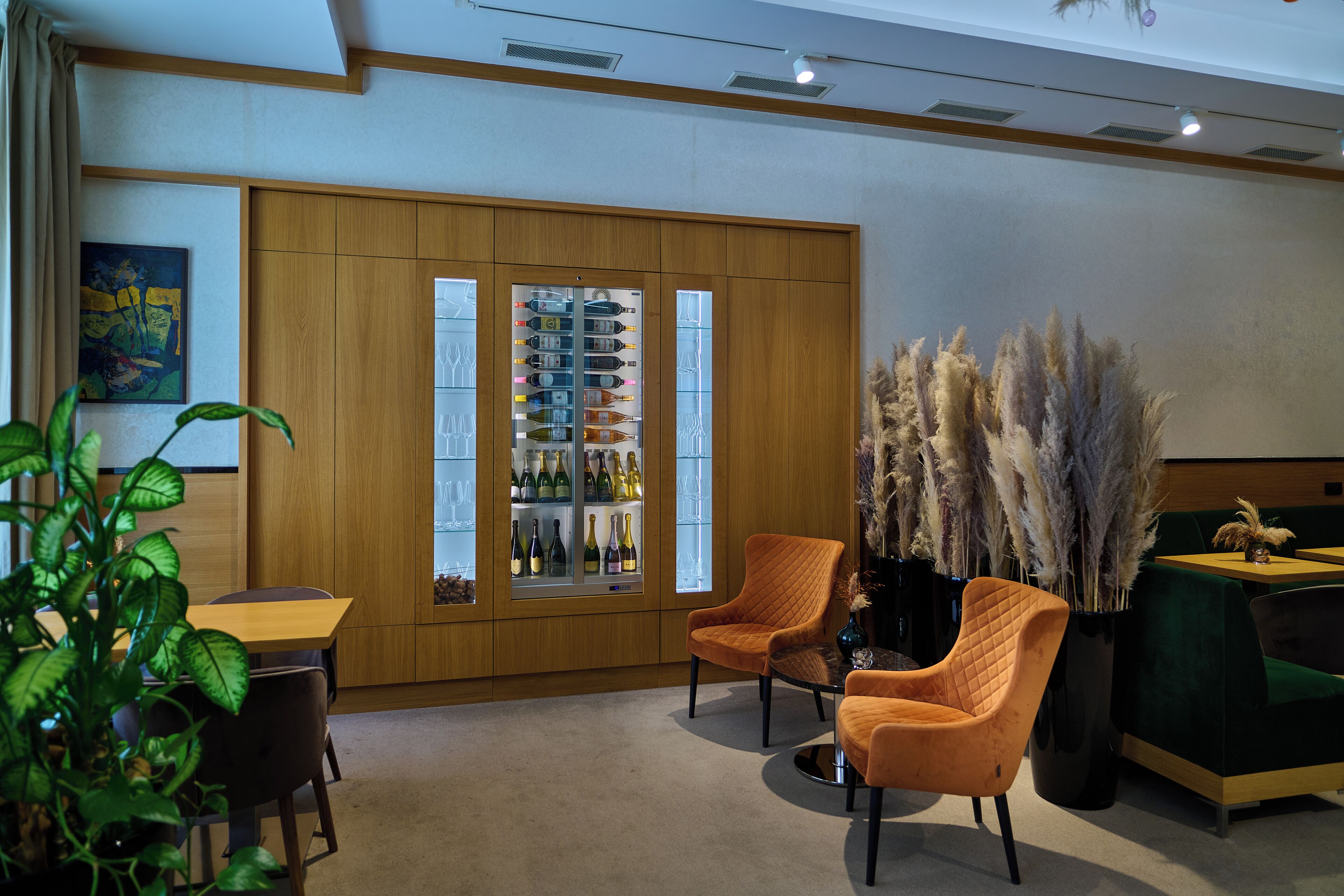 A modern interior with a wooden cabinet displaying wine bottles, two orange chairs, a green sofa, and a potted plant.
