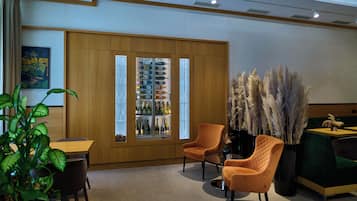 A modern interior with a wooden cabinet displaying wine bottles, two orange chairs, a green sofa, and a potted plant.
