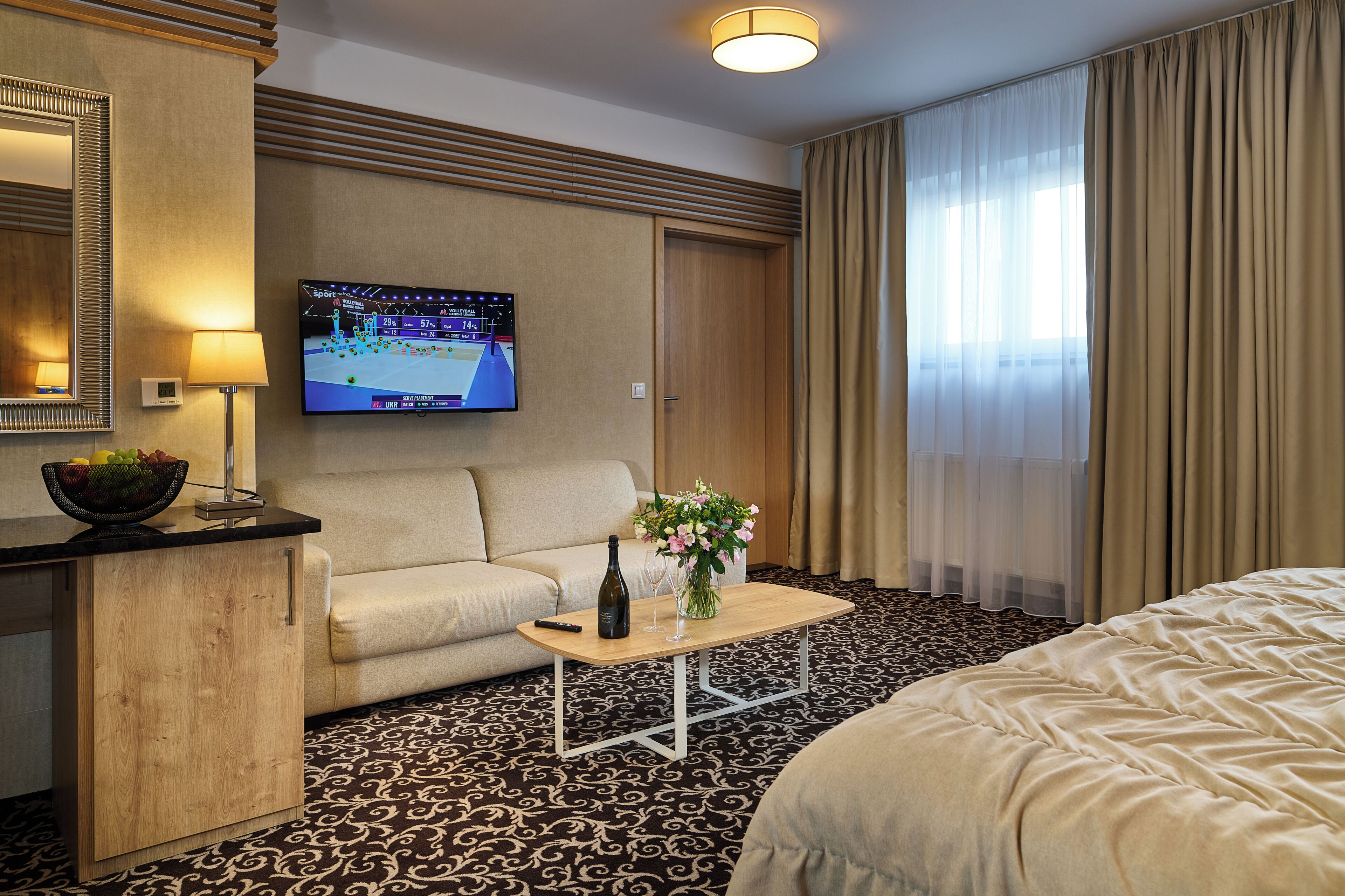A hotel room with a sofa, a coffee table with a bottle and flowers, a TV on the wall, and a bed.