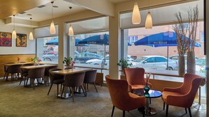 Interior de un restaurante con mesas, sillas, lámparas colgantes y ventanas amplias que dan vista a coches aparcados y un edificio.