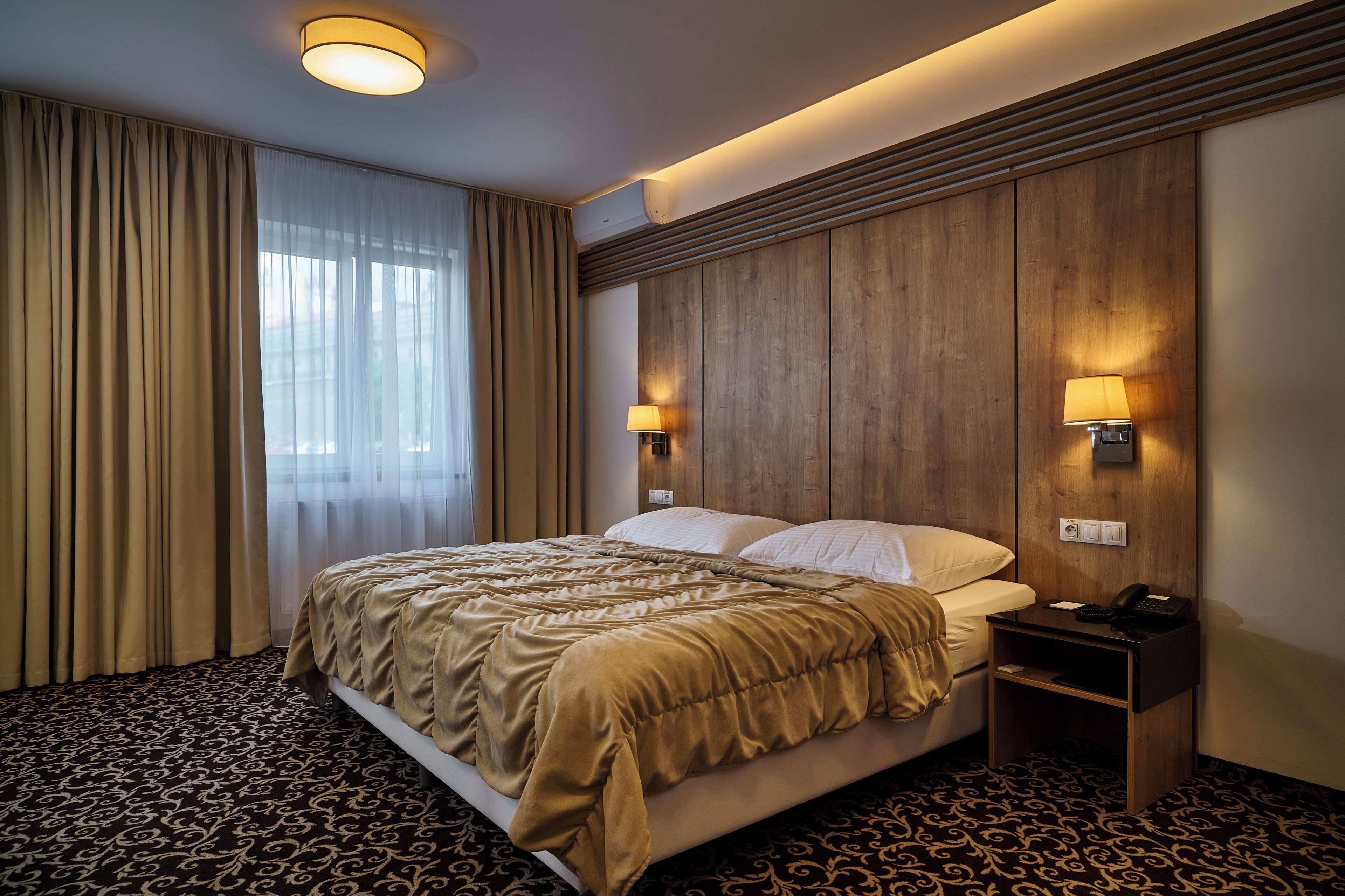 Camera d'albergo con un letto, tende, una testiera in legno, due lampade a parete e un comodino con il telefono.