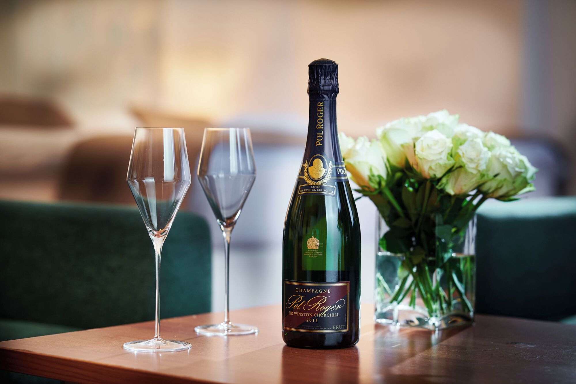 A bottle of Pol Roger champagne, two empty glasses, and a vase of white flowers on a wooden table.