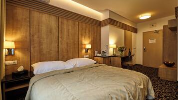 A hotel room with a large bed, wooden paneling, a desk with a lamp, a small table with a vase, and a door with a sign.