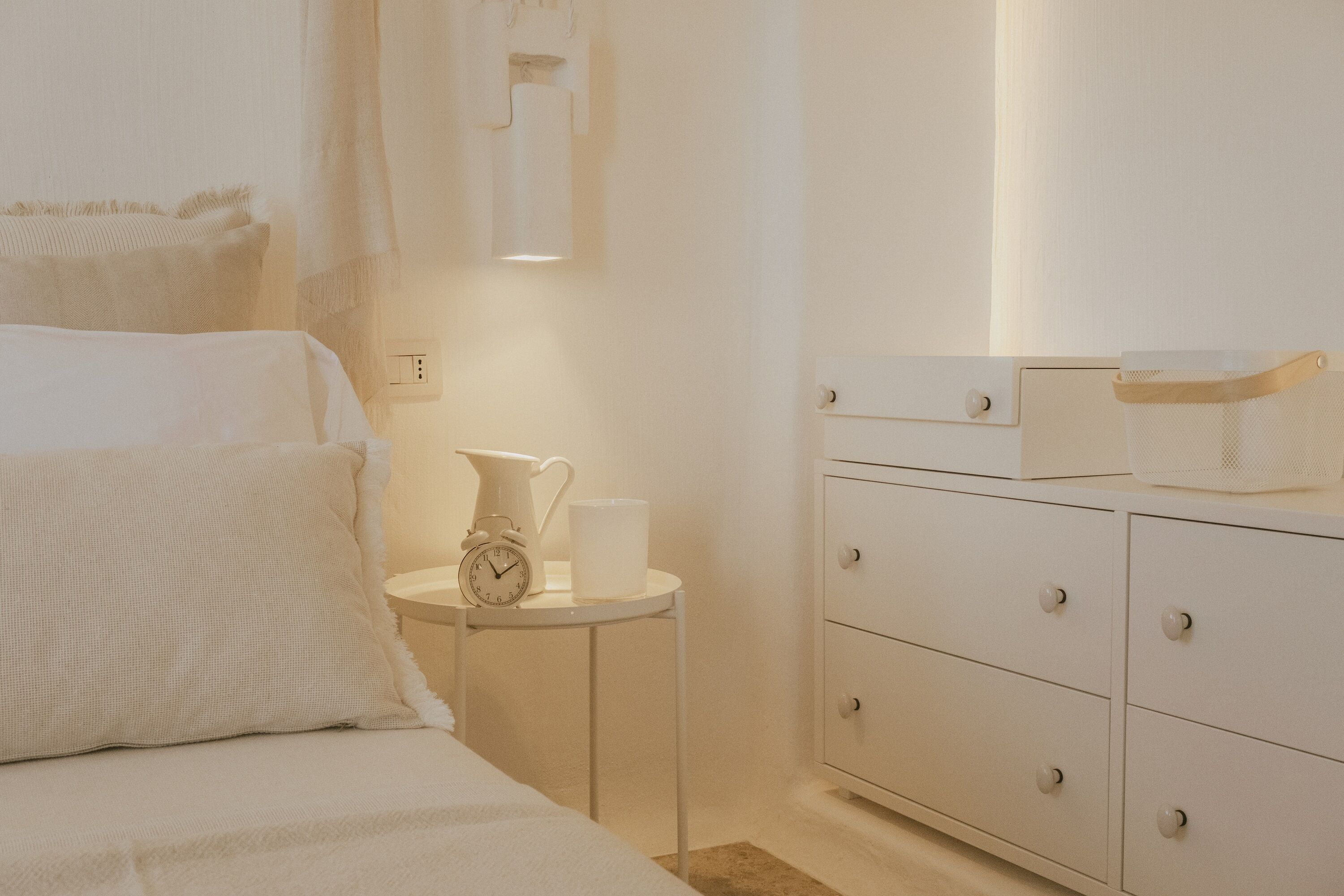 Un dormitorio con una cama, una mesita de noche con un reloj y una taza, y un aparador con cajones.