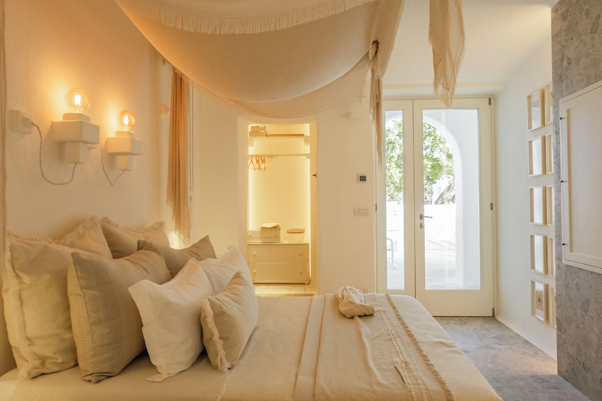 A bedroom with a large bed, a canopy, bedside lamps, a wardrobe, and a bathroom visible through an open door.