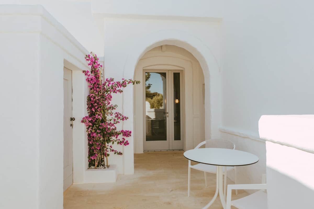 Ein heller Innenraum mit einer Tür, die auf einen Balkon führt, einem kleinen Tisch und einem Blumentopf mit rosa Blüten.