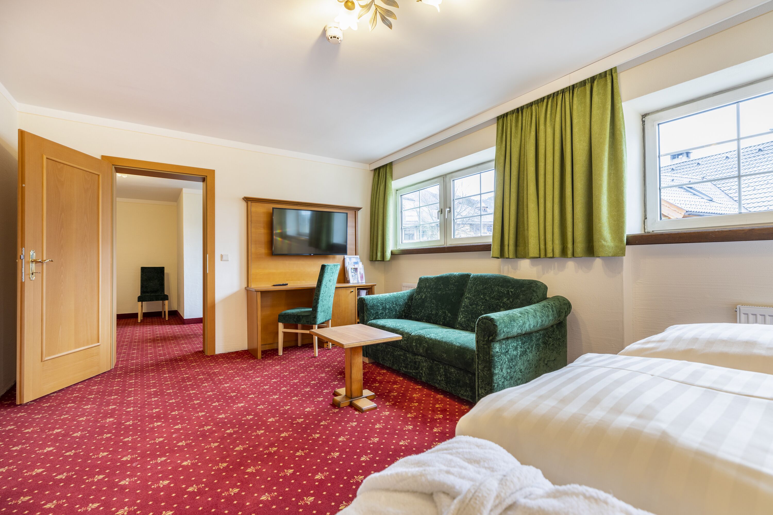 Une chambre d’hôtel avec un tapis rouge, un canapé vert, un bureau en bois, une télévision et une fenêtre avec des rideaux verts.