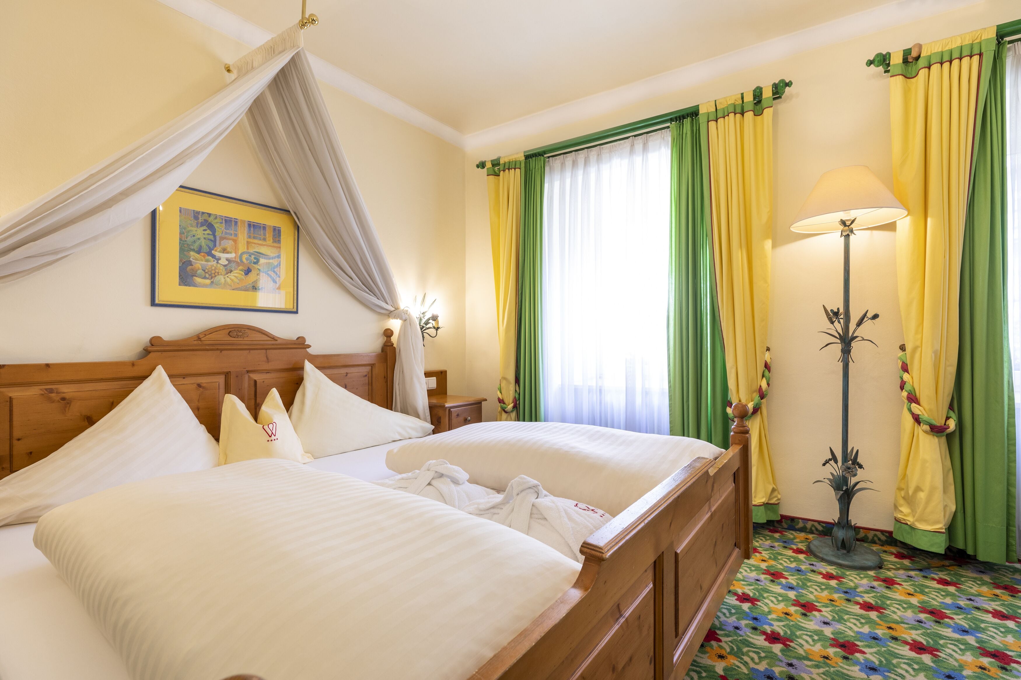 A double bed with white linens, a wooden headboard, and a canopy. There is a bedside table with a lamp, a framed picture on the wall, and a colorful patterned carpet.