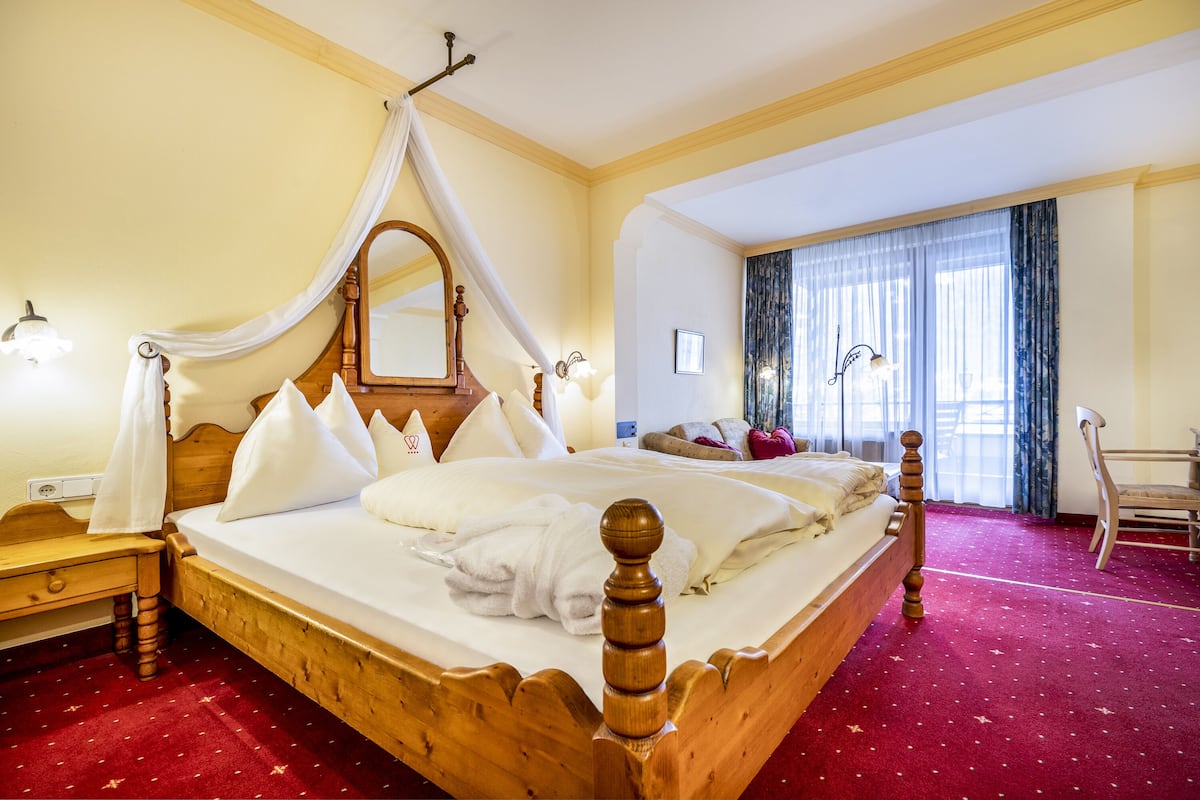 Ein Hotelzimmer mit einem hölzernen Bett, einem Baldachin, einem roten Teppich und einem großen Fenster mit Vorhängen.