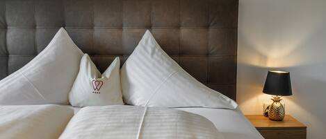 Una cama bien hecha con sábanas blancas y dos almohadas, un cabecero marrón oscuro y una lámpara de noche sobre una mesita de madera.