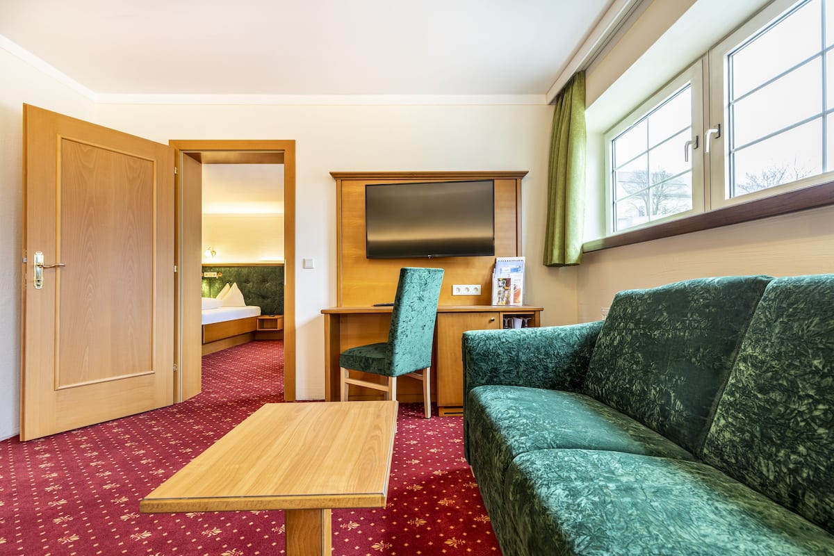 Ein Hotelzimmer mit Holztür, einem grünen Sofa, einem Holztisch, einem an der Wand montierten Fernseher und einem Fenster mit Vorhängen.