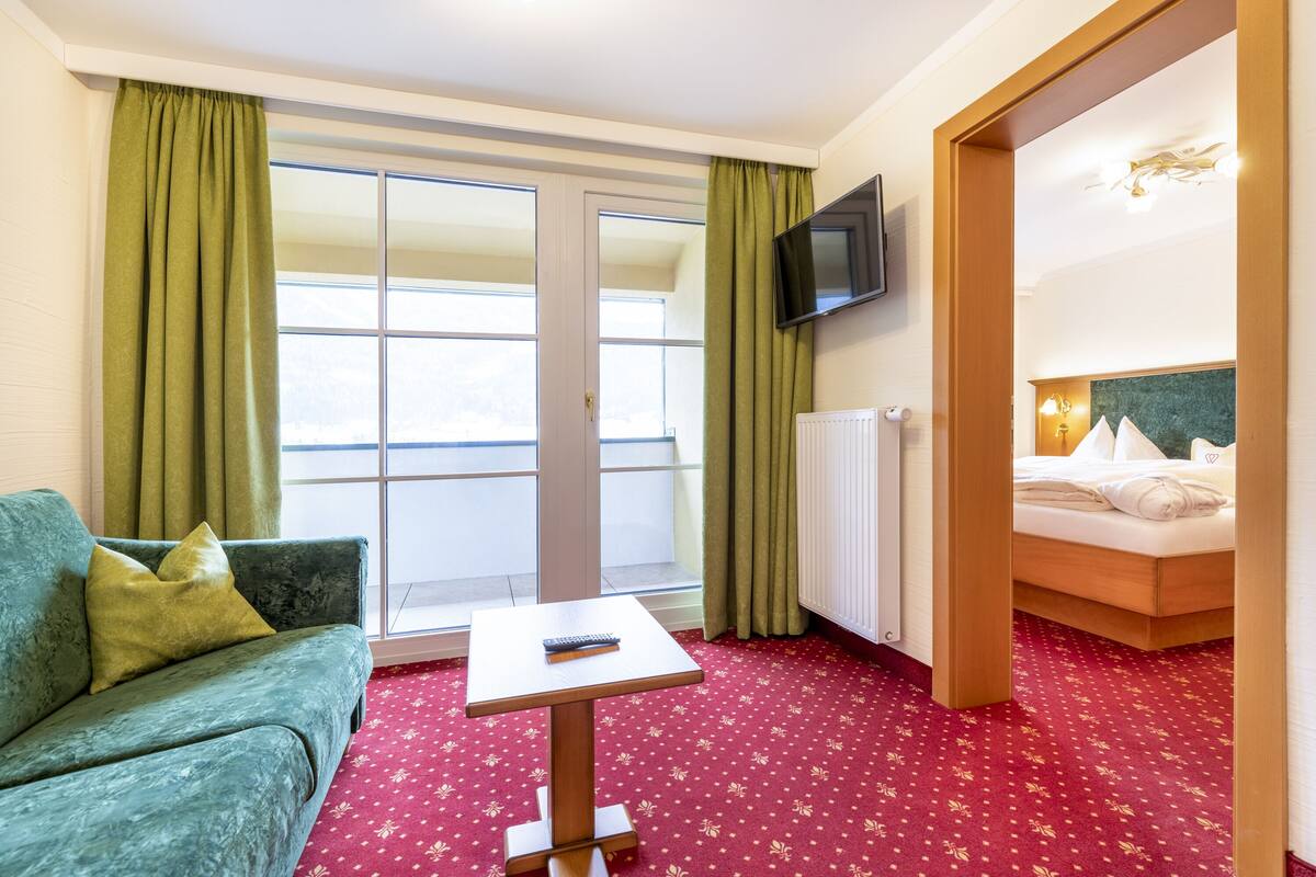 Ein Hotelzimmer mit einem grünen Sofa, einem kleinen Tisch, einem Bett mit weißen Leinen und einem an der Wand montierten Fernseher sowie einem roten Teppich mit Muster.