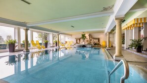 Ein großes Hallenbad mit klarem, blauem Wasser, gelben Liegestühlen und Pflanzen rund um den Poolbereich.