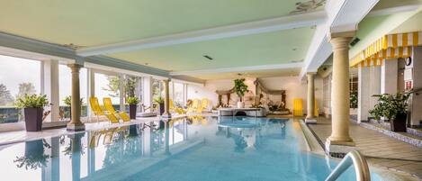 A large indoor swimming pool with clear blue water, yellow lounge chairs, and potted plants around the pool area.