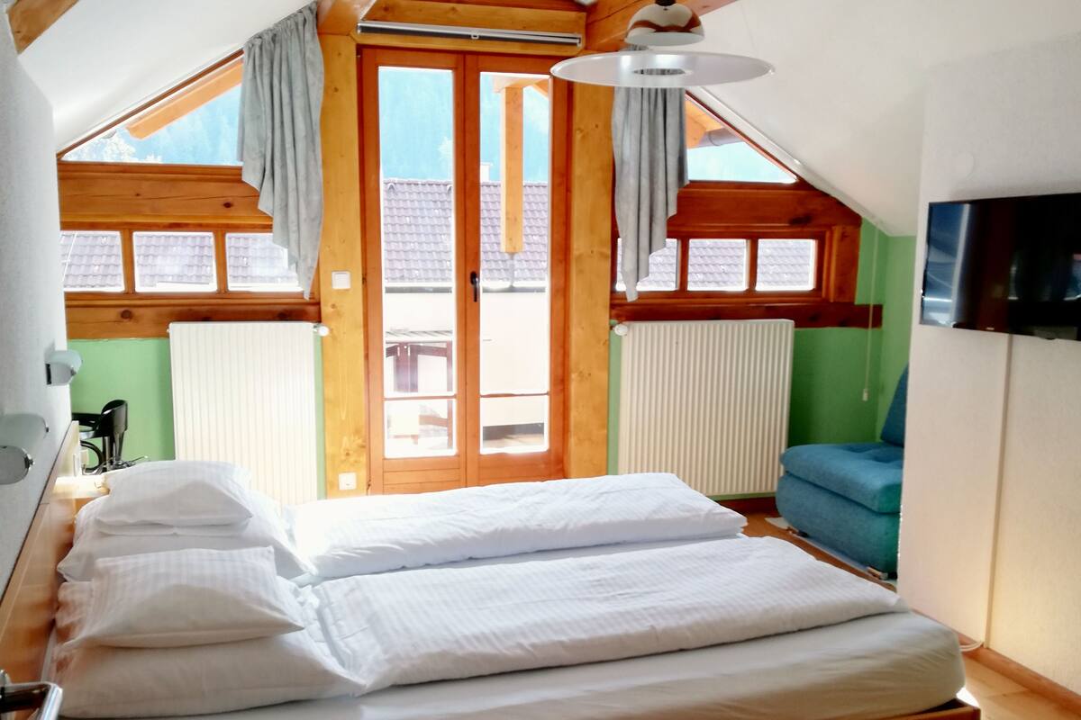 Un dormitorio con techo de madera, cama con ropa de cama blanca, televisor en la pared y vistas al exterior por una ventana.