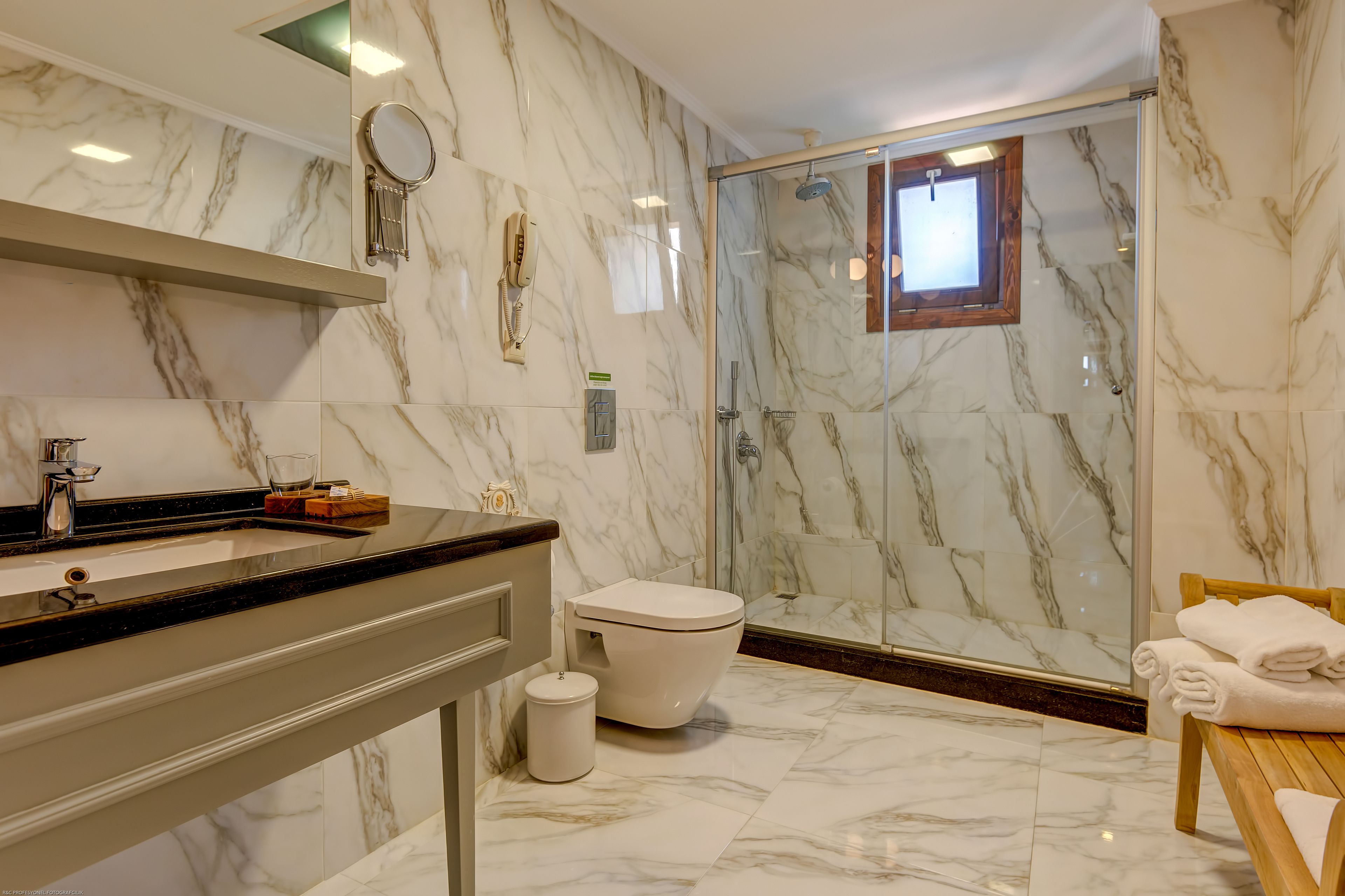 Un baño moderno con suelo de mármol, ducha con cerramiento de vidrio, inodoro y tocador con espejo y teléfono.