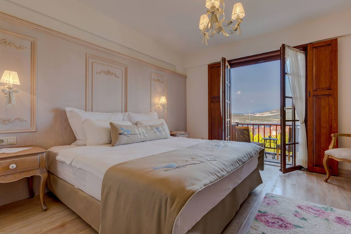 A bedroom with a large bed, bedside lamps, a wooden nightstand, a chair, and a balcony with a view.