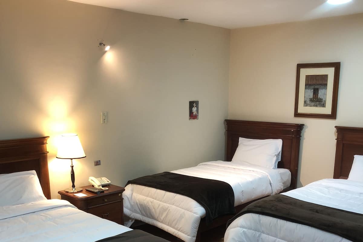 Habitación de hotel con tres camas, una mesita de noche con lámpara, teléfono y un cuadro en la pared.