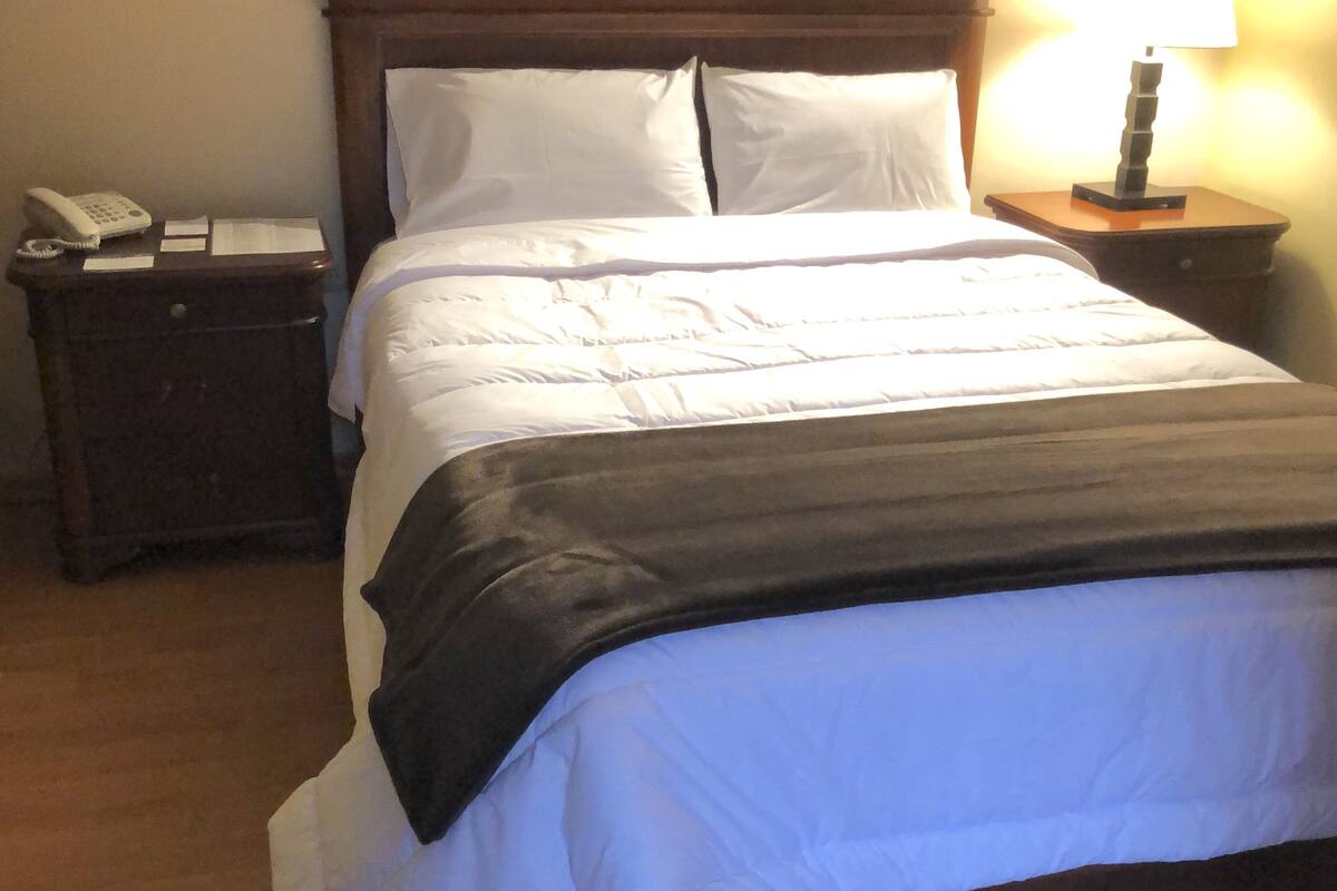 Una cama bien hecha con sábanas blancas y una manta oscura, una mesita de noche de madera con una lámpara y un teléfono a la izquierda.