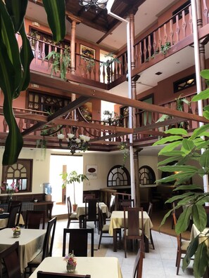 Interior de un hotel de varios niveles con balcones de madera, plantas de interior y un comedor con mesas y sillas.