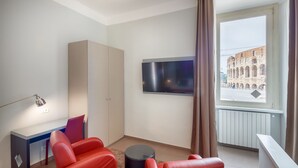 Une chambre moderne équipée d’un bureau, de deux chaises rouges, d’une table ronde, d’un téléviseur et offrant une vue sur le Colisée par la fenêtre.