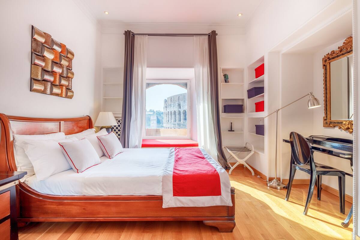 A hotel room with a large bed, a desk, a chair, a bookshelf, and a view of a historic building.