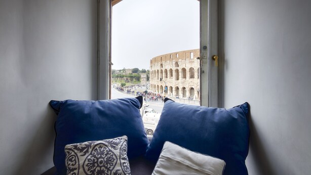Une pièce avec vue sur le Colisée, visible à travers une fenêtre, comprenant deux coussins bleus et un coussin à motifs sur un canapé de couleur foncée.